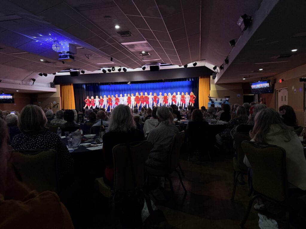 Audience watches a group of performers in red costumes on a stage under blue lighting in a hall.