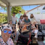 Smiling family gathers on a sunny backyard patio, posing for a selfie with kids, an elderly person, and several adults in the background.