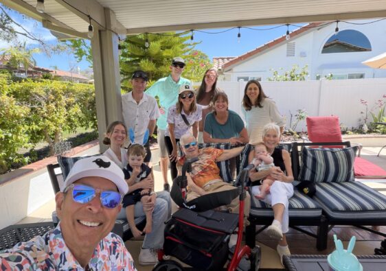Smiling family gathers on a sunny backyard patio, posing for a selfie with kids, an elderly person, and several adults in the background.