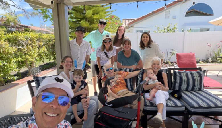 Smiling family gathers on a sunny backyard patio, posing for a selfie with kids, an elderly person, and several adults in the background.