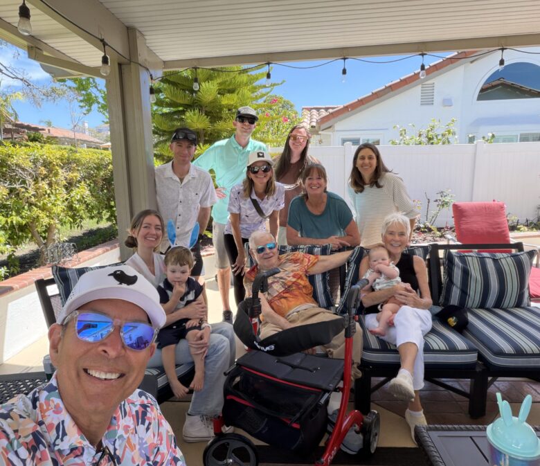 Smiling family gathers on a sunny backyard patio, posing for a selfie with kids, an elderly person, and several adults in the background.