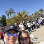 Two people smiling for a selfie at an outdoor event, with a line of attendees and tropical plants in bright sun.
