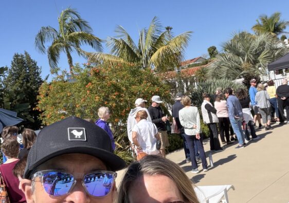Two people smiling for a selfie at an outdoor event, with a line of attendees and tropical plants in bright sun.