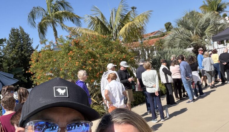 Two people smiling for a selfie at an outdoor event, with a line of attendees and tropical plants in bright sun.