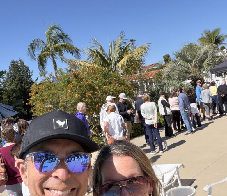 Two people smiling for a selfie at an outdoor event, with a line of attendees and tropical plants in bright sun.