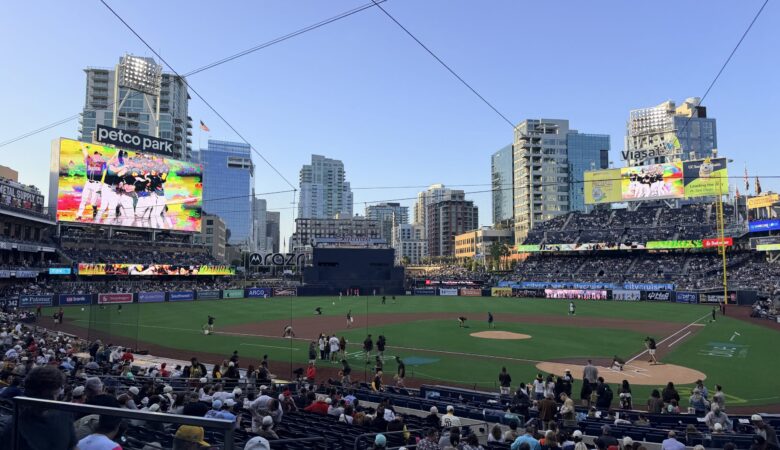 A packed baseball stadium with players on the infield and a large video board displaying colorful graphics; 'petco park' sign visible at left.